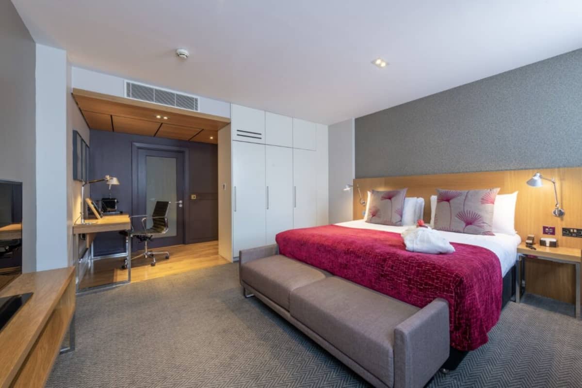A modern hotel room with a large bed, a desk, a chair, and a bench.