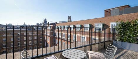 A balcony with a view of a cityscape, including a church spire, and furnished with two chairs and a small table.