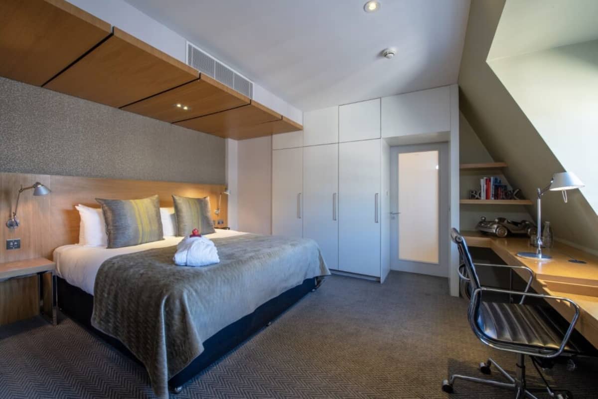 A modern hotel room with a large bed, a desk, and a chair. There is a wardrobe and a door leading to another room.