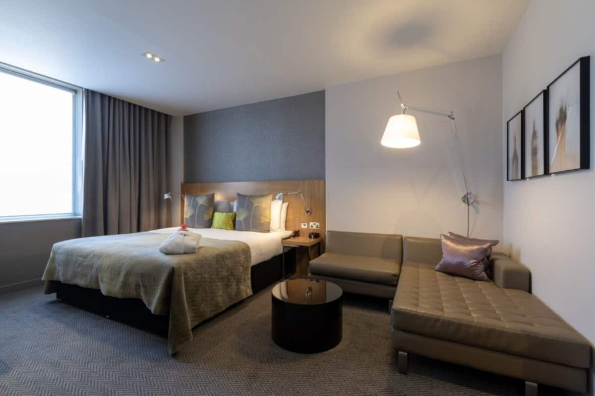 A hotel room with a bed, a sofa, a round coffee table, and a desk with a lamp.