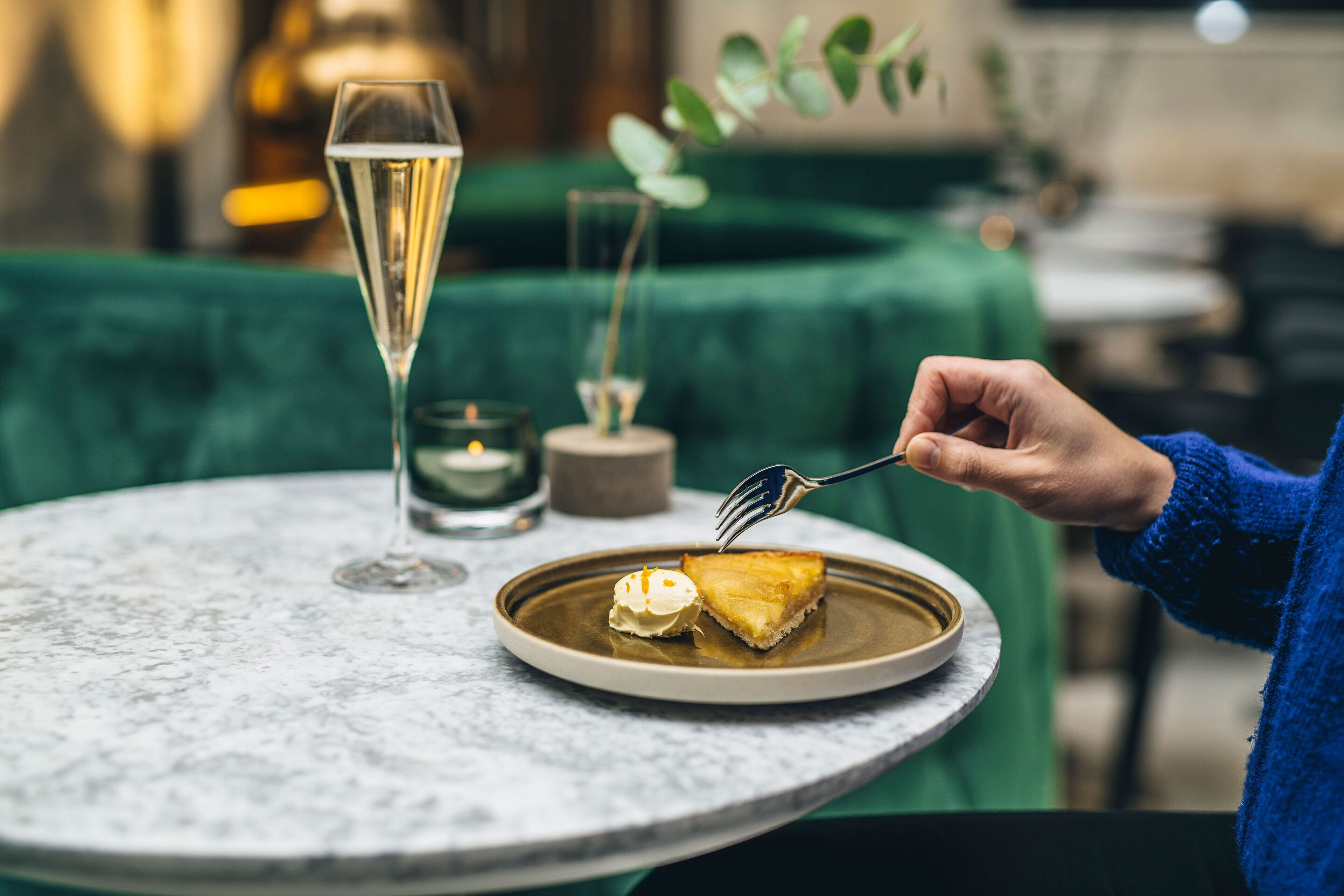 Een persoon serveert een plak gebak op een bord, met een vork, een glas champagne en een kaarsje op een tafel.