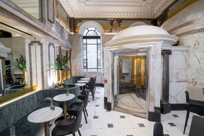 Un hall d’entrée luxueux, avec un sol en marbre, un grand escalier et un espace salon agrémenté de tables et de chaises rondes.