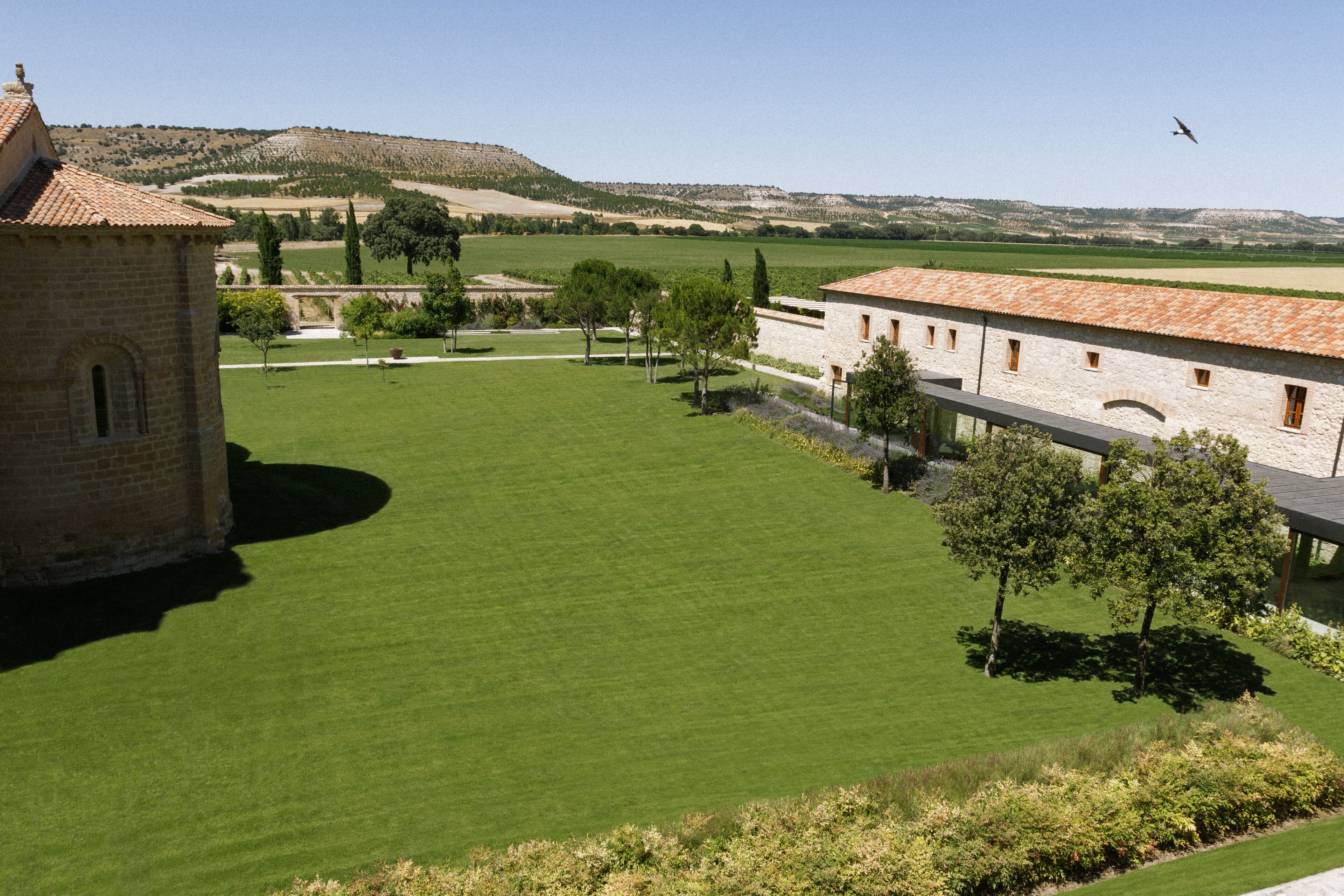 Un patio con un edificio de techo con tejas y paredes de piedra, rodeado de césped verde y árboles.