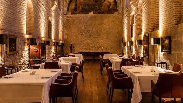 Une salle à manger avec de hauts plafonds en voûte et des murs en briques. Les tables sont dressées avec des nappes blanches et des chaises rouges.