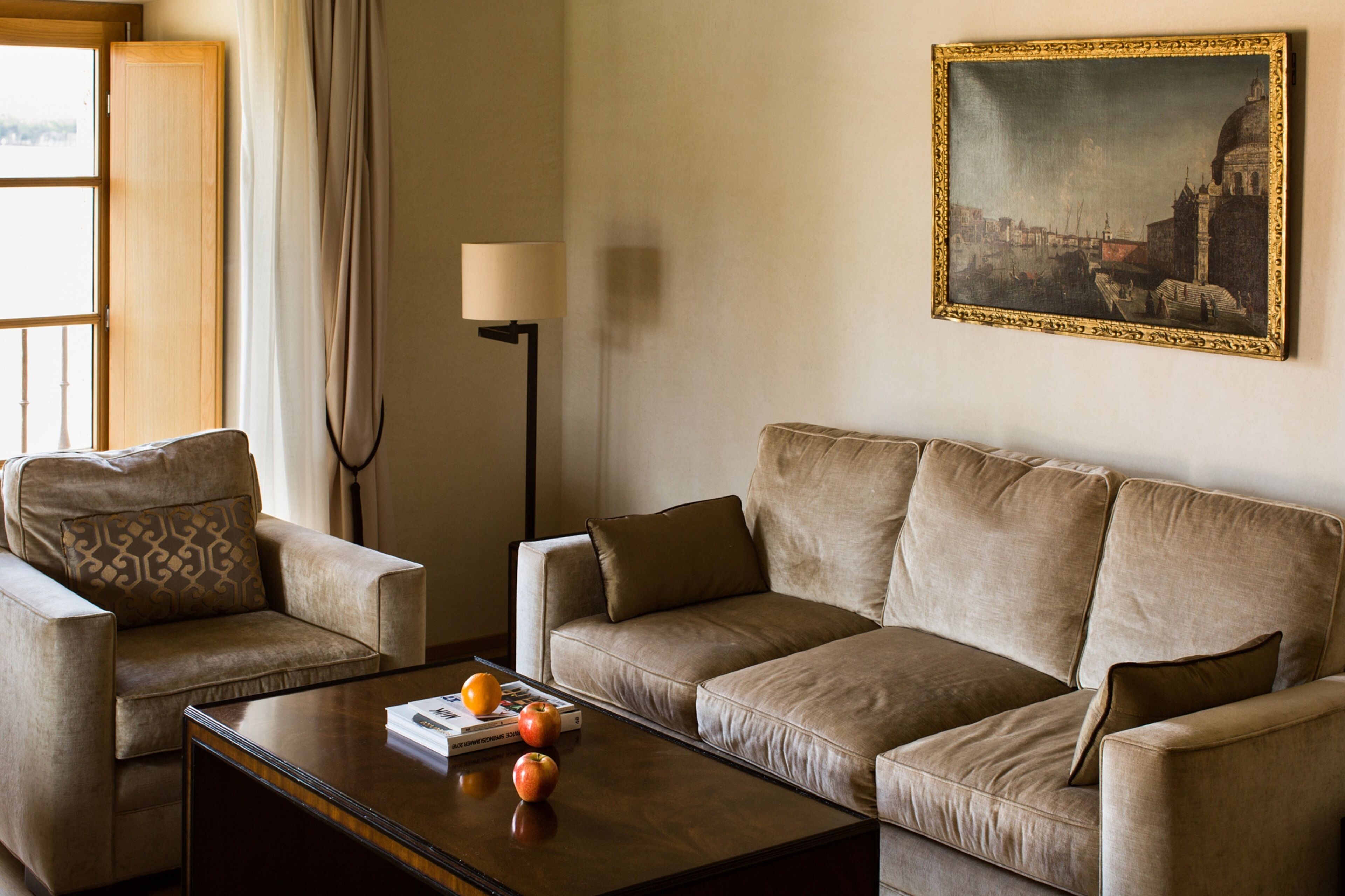 Una sala de estar con un sofá color beige, un sillón a juego y una mesa de centro con libros y naranjas. Una lámpara y un cuadro enmarcado están en la pared.