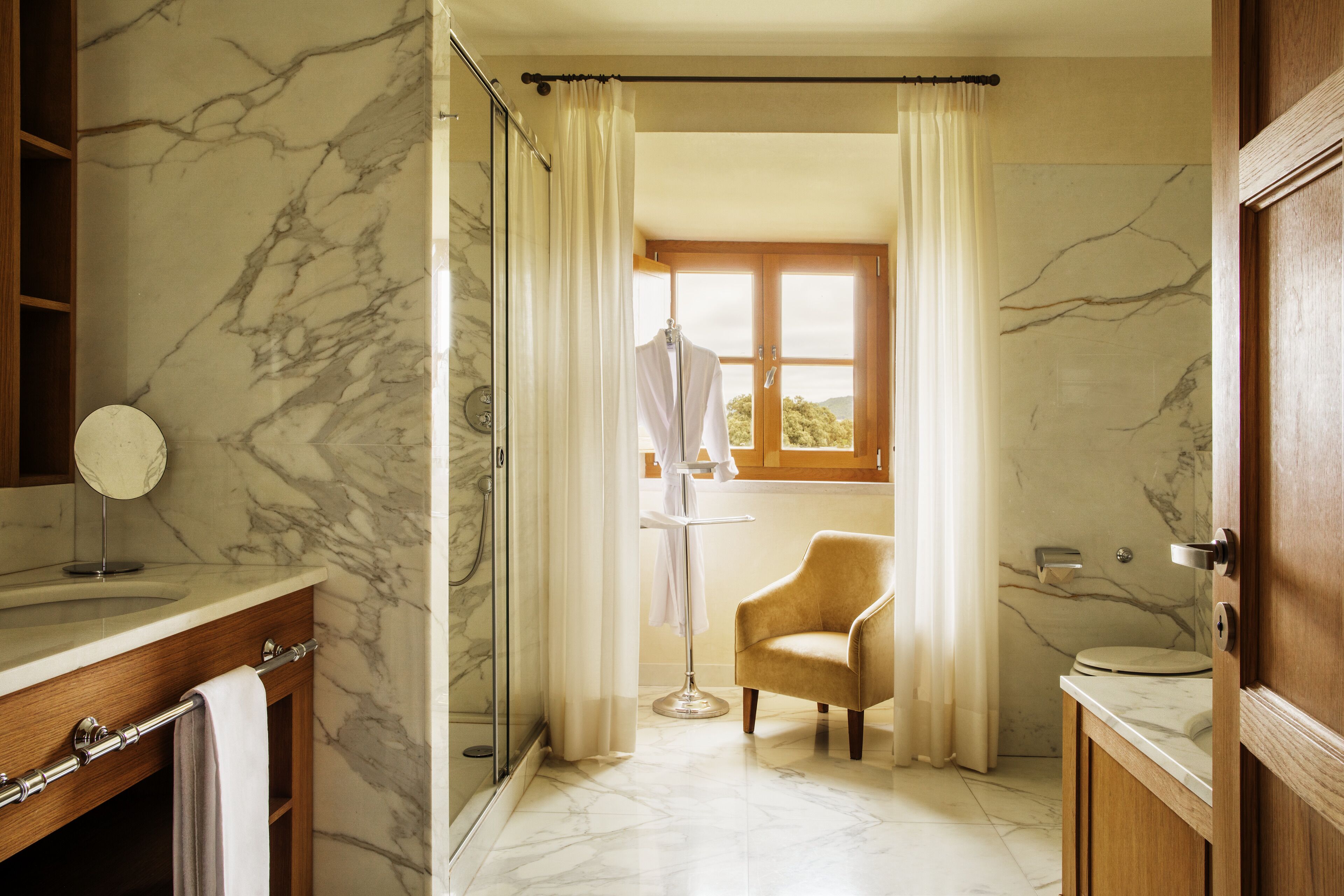 Un baño con pared de mármol, tocador de madera, una silla y una ventana con cortinas.