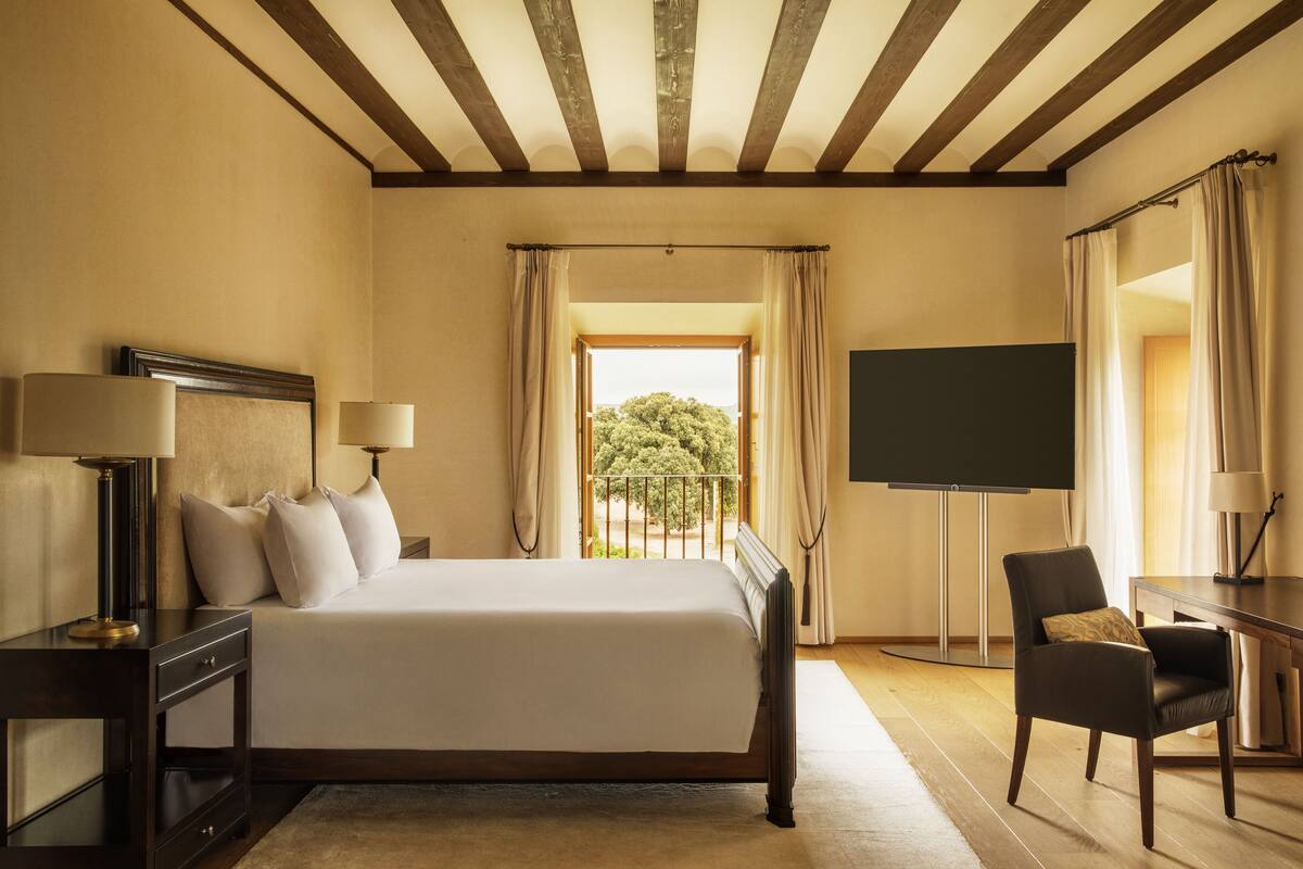 Ein Hotelzimmer mit einem großen Bett, einem Schreibtisch mit Stuhl, einem Flachbildfernseher und einem Balkon mit Vorhängen.