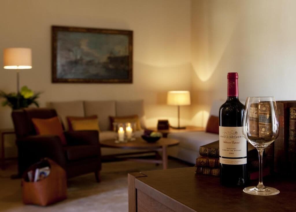 Habitación de hotel con cama, sofá, mesa de centro y una botella de vino con una copa.