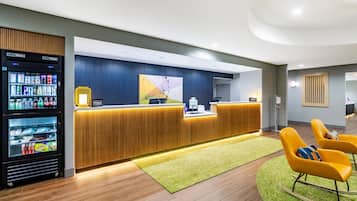 A hotel reception area with a wooden reception desk, a minibar, and a seating area with yellow chairs.
