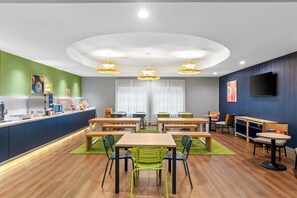 A modern dining area with wooden tables and chairs, a counter with a coffee machine, and a large window with curtains. - Spark by Hilton Missouri City Sugar Land (Missouri City)