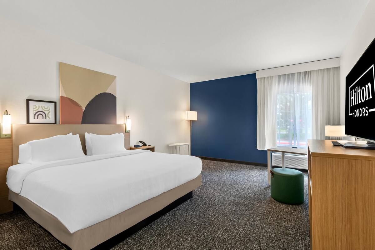 Une chambre d’hôtel avec un lit, un bureau, un pouf vert, une grande fenêtre avec des rideaux et un téléviseur fixé au mur.