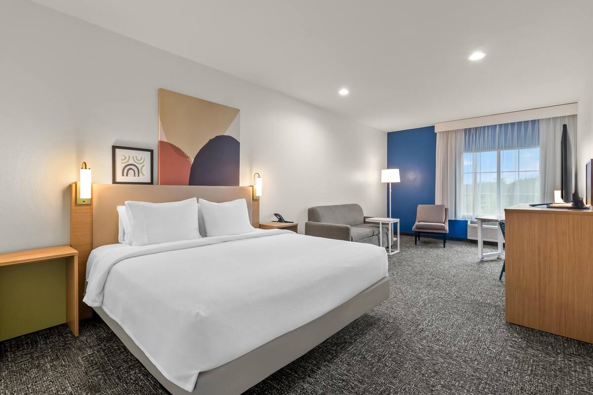 Une chambre d’hôtel comprenant un lit, un bureau, une chaise, une lampe de bureau, un téléviseur fixé au mur et une fenêtre avec des rideaux.