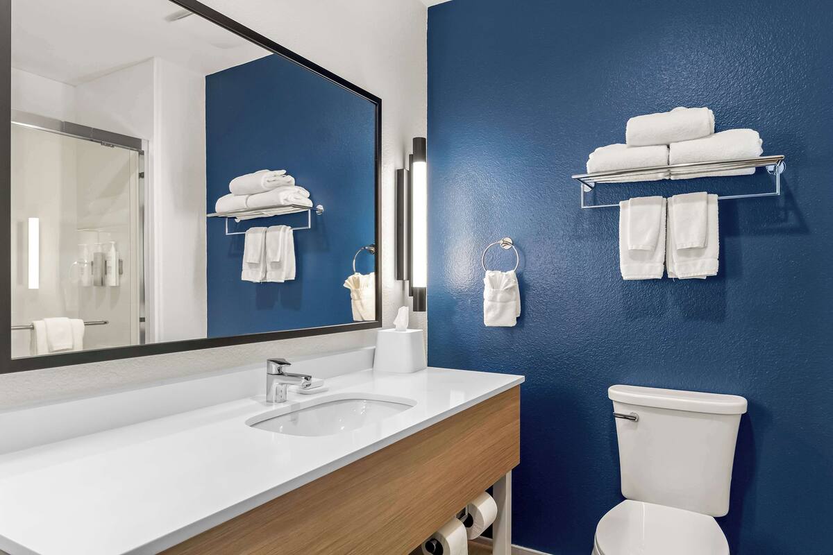 Une salle de bain moderne avec un lavabo blanc, un grand miroir, un mur d’accent bleu et des serviettes rangées avec soin sur des crochets.
