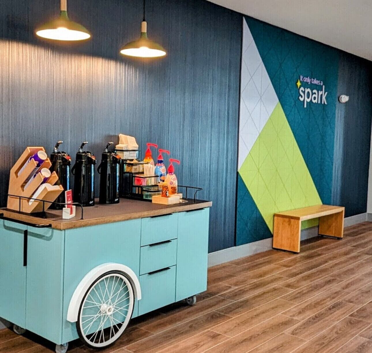 A room with a teal-colored cart with a wheel, a wooden bench, and a wall with a geometric design and the text "Only takes a spark".