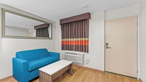 A room with a blue sofa, a white table, a mirror, a window with striped curtains, a wall-mounted air conditioner, and a door. - Guest Inn (Rogers)