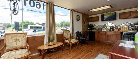 A spacious room with wooden flooring, a large window with curtains, a television mounted on the wall, and a kitchen area with wooden cabinets and a countertop.