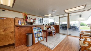 A reception area with a wooden counter, a trash bin, a desk with a computer, and a window offering a view of the street. - Guest Inn (Rogers)
