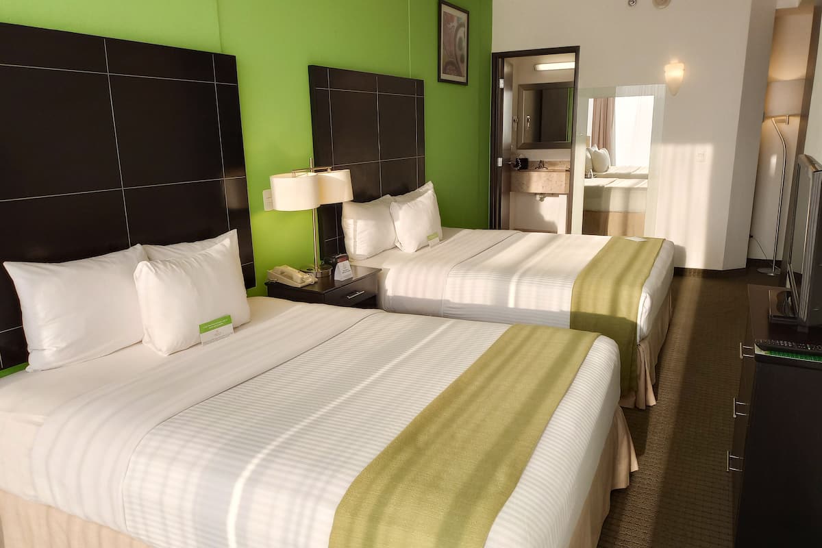 Une chambre d’hôtel avec deux lits, un mur d’accent de couleur verte, une salle de bain avec un miroir et un téléviseur.