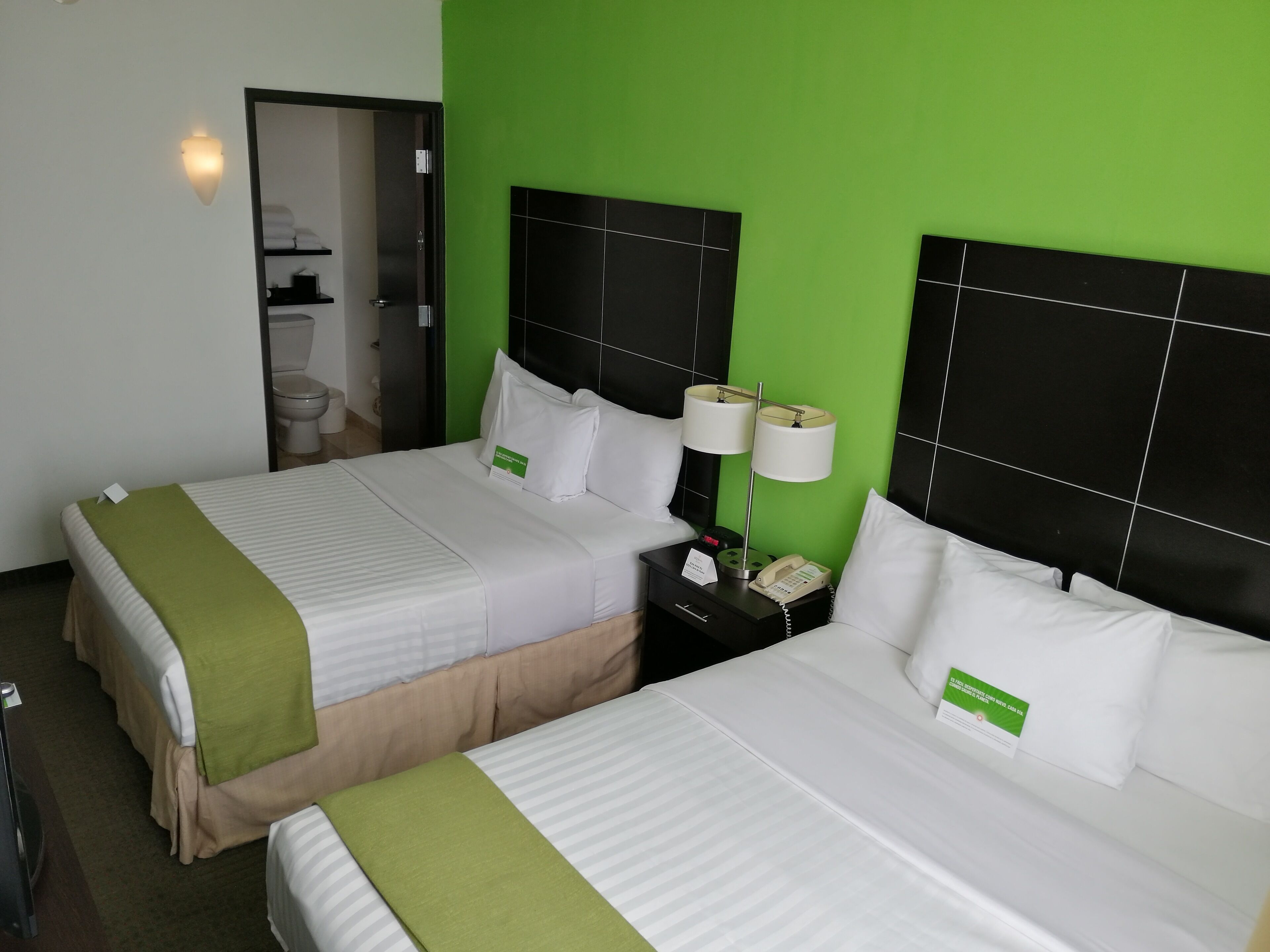 Ein Hotelzimmer mit zwei Betten, jeweils mit weißer Bettwäsche und grünen Zierkissen. An den Nachttischen stehen Lampen, ein Telefon und ein Schild. Der Raum hat eine grüne Akzentwand und ein schwarzes Kopfteil.