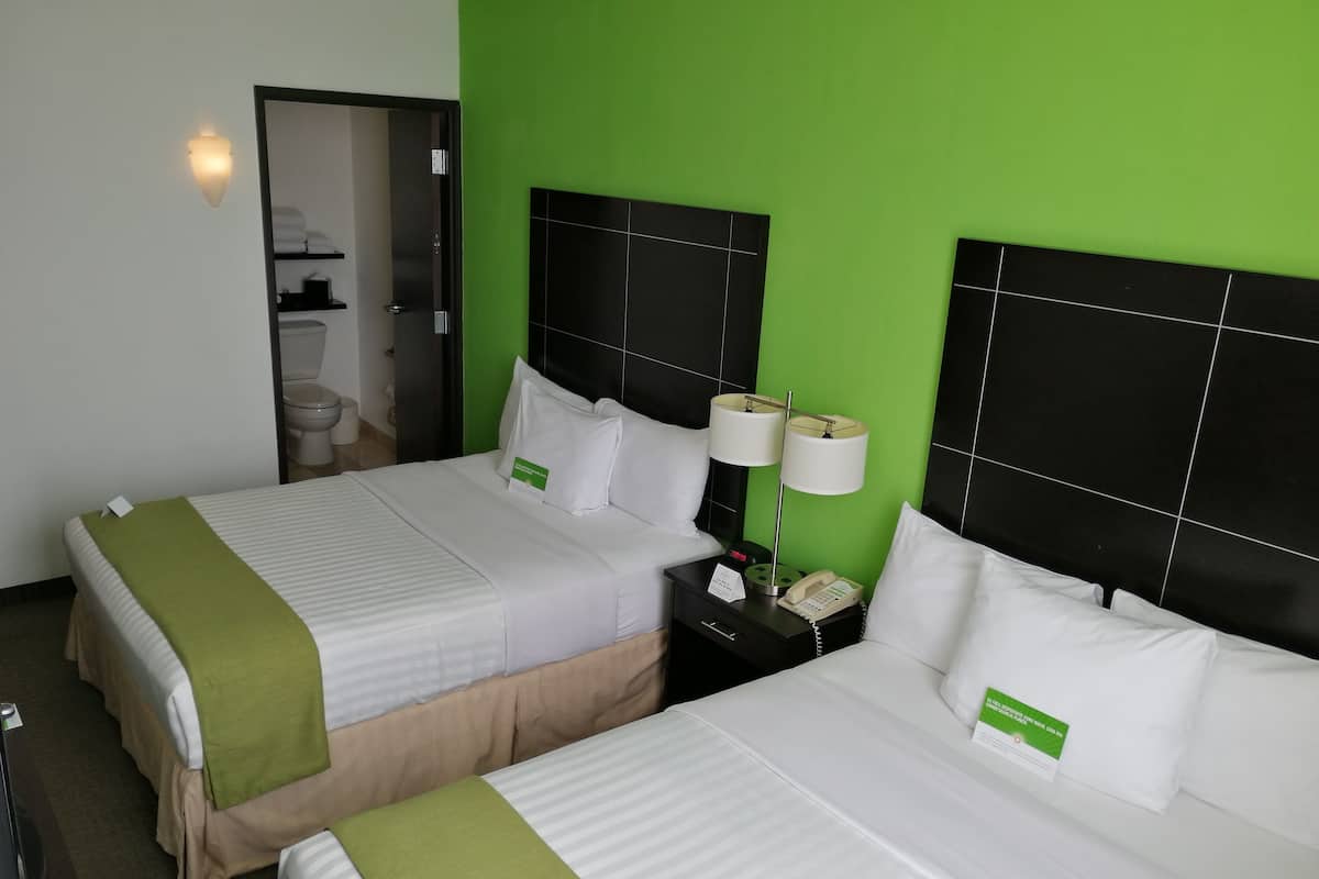 Une chambre d’hôtel avec deux lits, chacun agrémenté de draps blancs et de coussins verts. On y trouve une table de chevet avec une lampe, un téléphone et un panneau. La chambre est dotée d’un mur d’accent vert et d’une tête de lit noire.