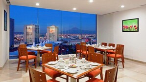 Un comedor con mesas y sillas, un ventanal con vistas a la ciudad y un cuadro en la pared.