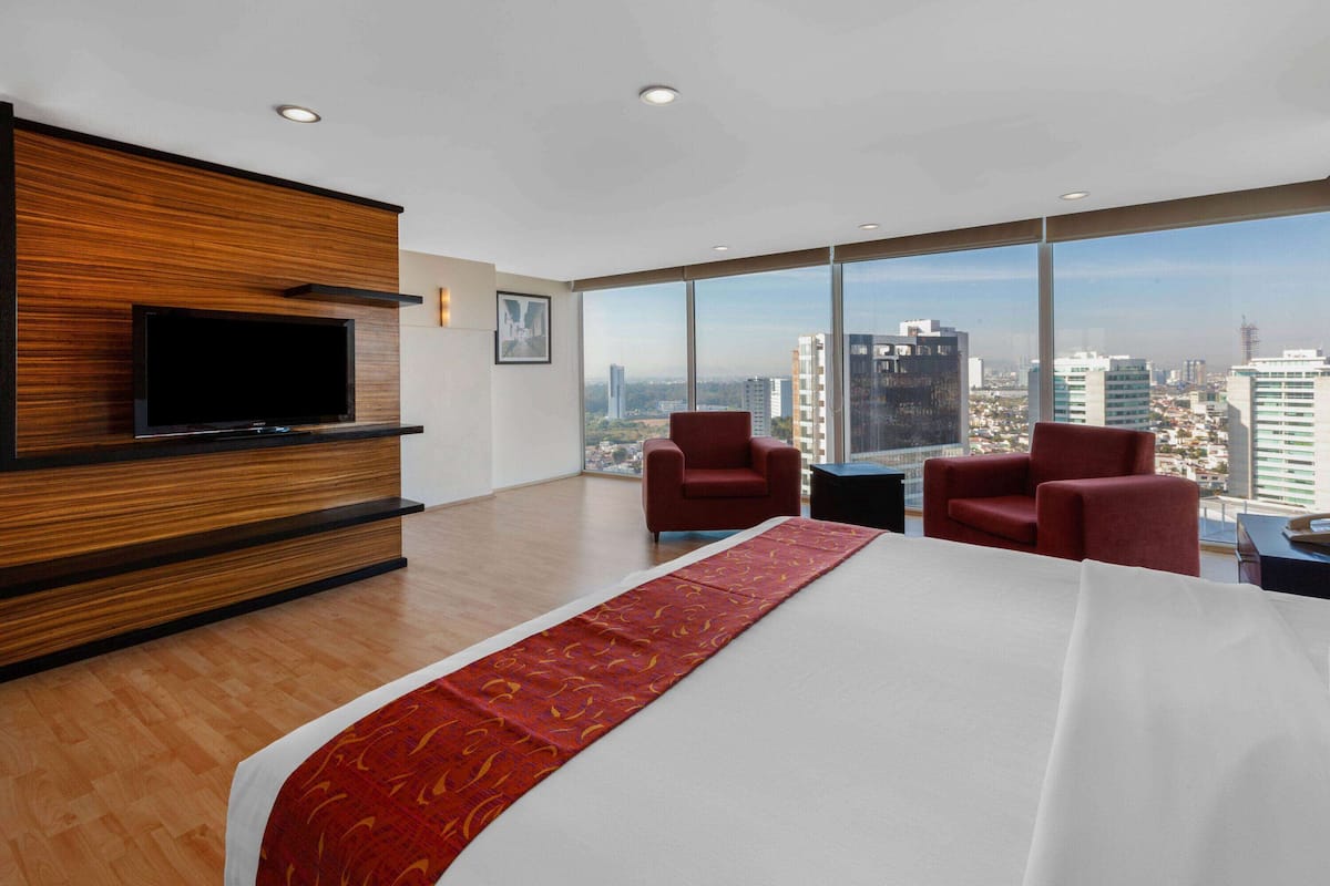 Une chambre d’hôtel avec un grand lit, deux fauteuils rouges, un téléviseur à écran plat fixé sur un panneau en bois, et une vue sur la ville à travers de grandes fenêtres.