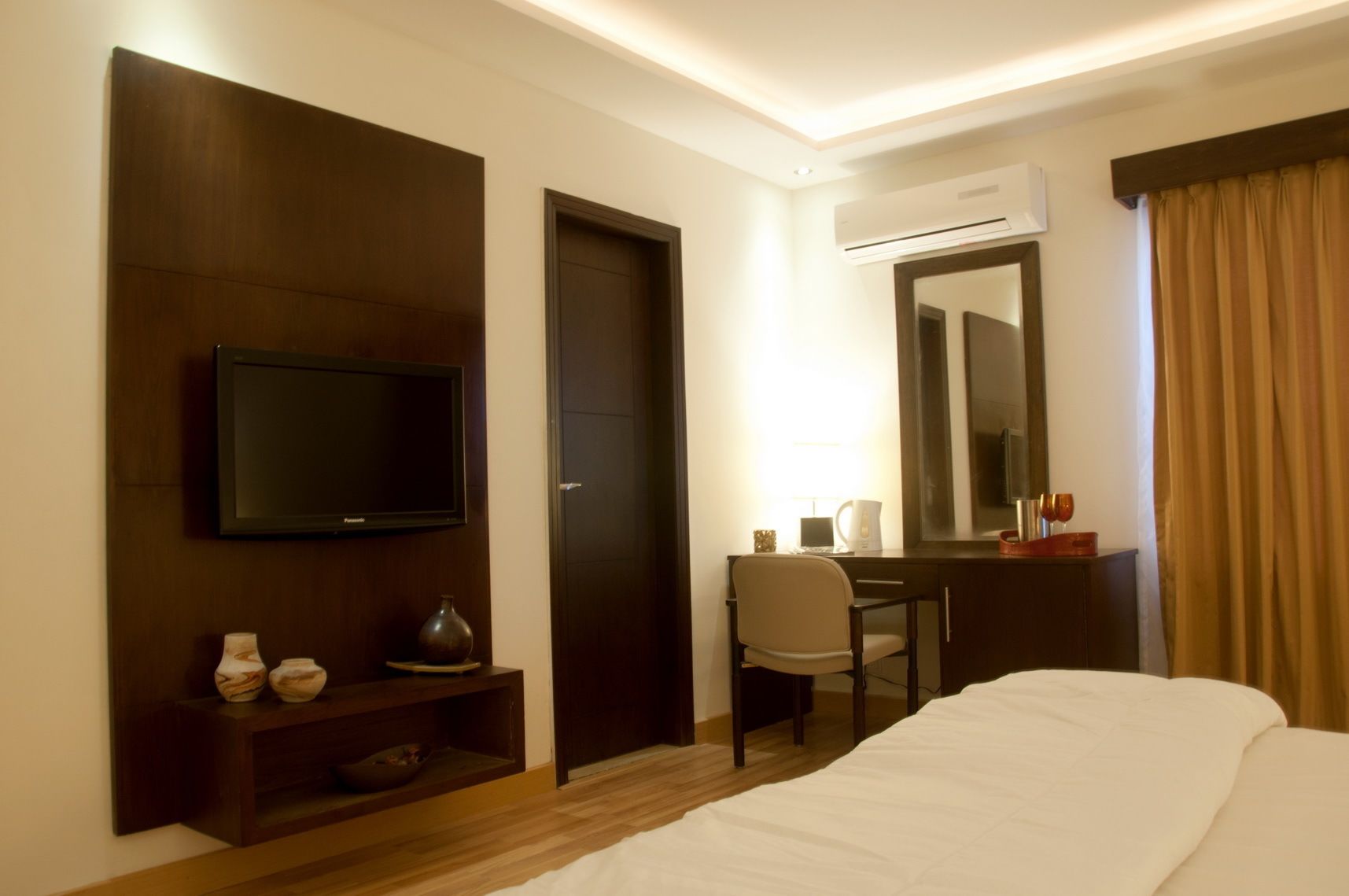 Une chambre d’hôtel comprenant un lit, un bureau avec une chaise, une télévision, une table de chevet agrémentée d’objets décoratifs, un miroir et un appareil de climatisation.