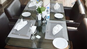 Zona pranzo con tavolo in vetro apparecchiata per quattro persone, circondata da poltrone in pelle. Sono presenti piante in vaso e vista sulla città attraverso ampie finestre.