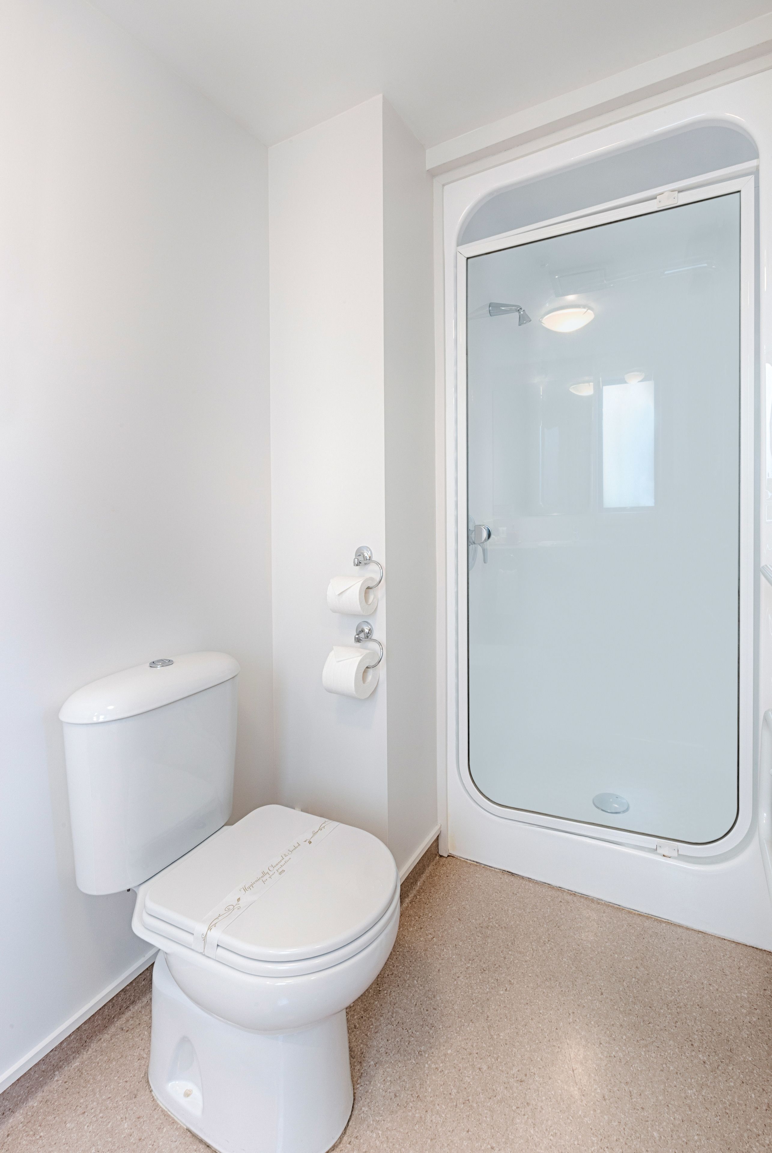 Un inodoro blanco con tapa, un box de ducha de vidrio y un dispensador de papel higiénico adherido a la pared.