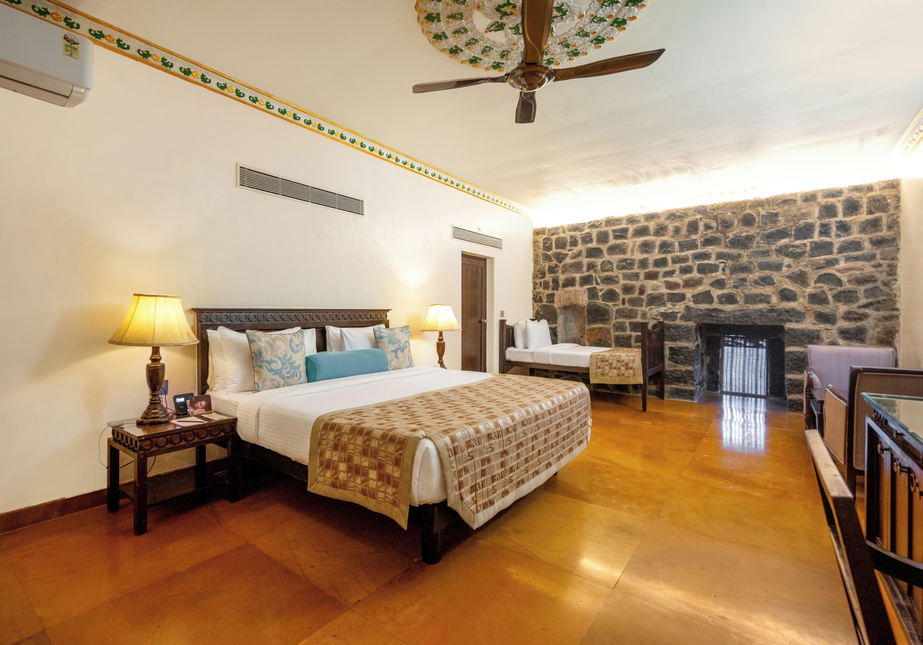 A bedroom with a bed, bedside lamps, a stone wall, a fireplace, and a ceiling fan.