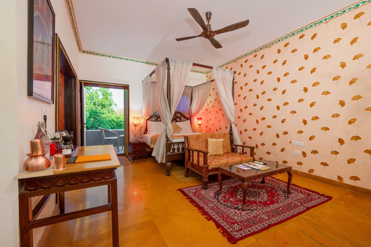Une pièce comprenant un lit, une table en bois, un tapis rouge à motifs, un ventilateur de plafond et un mur orné de motifs de feuilles.