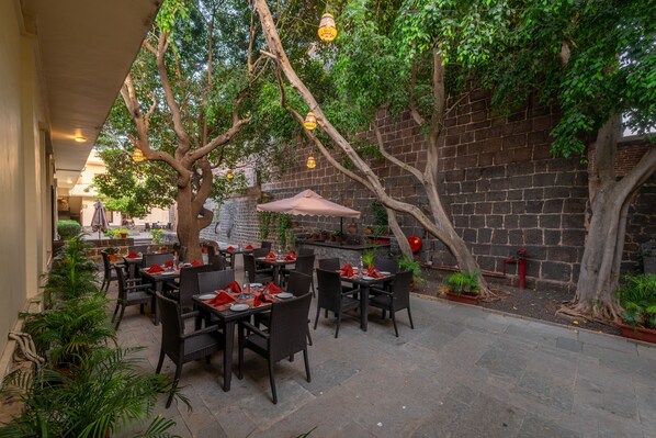 Un espace de restauration en plein air, avec des tables et des chaises, entouré d’arbres et d’un mur de briques.