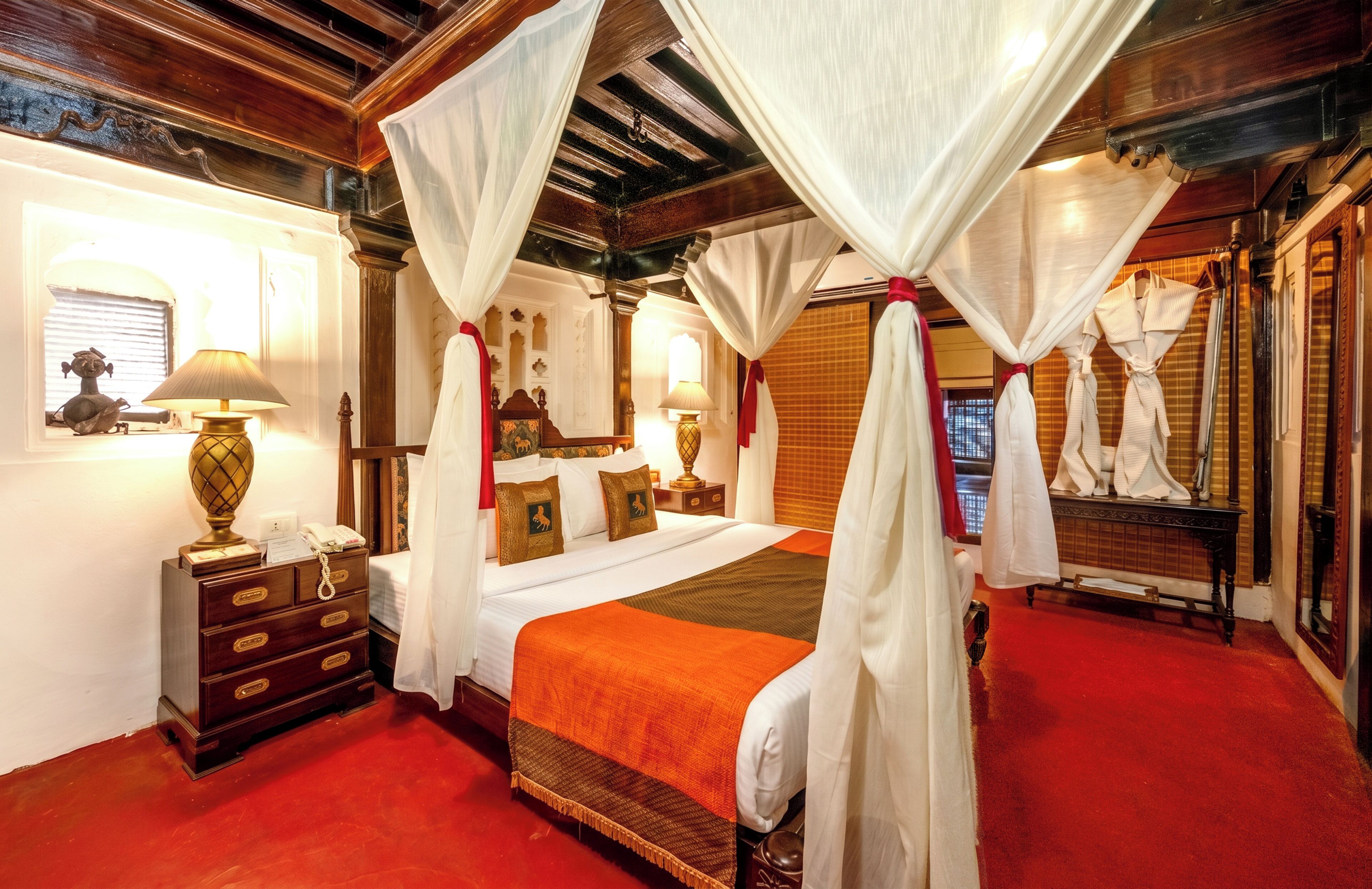 A four-poster bed with a canopy, a wooden nightstand with a lamp, a dresser with drawers, and a red carpeted floor.
