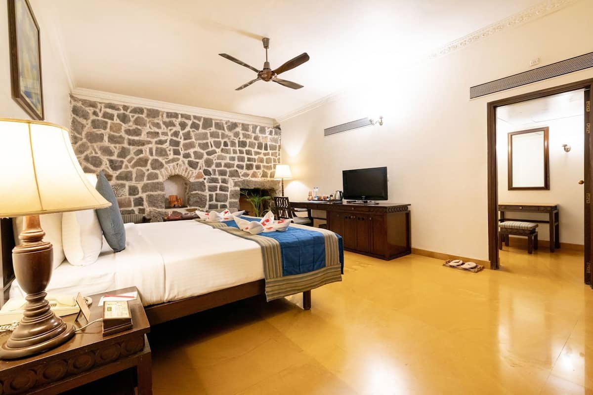 Une chambre d’hôtel avec un mur en pierre, un lit recouvert d’une courtepointe bleue, un bureau avec une télévision et un ventilateur de plafond.
