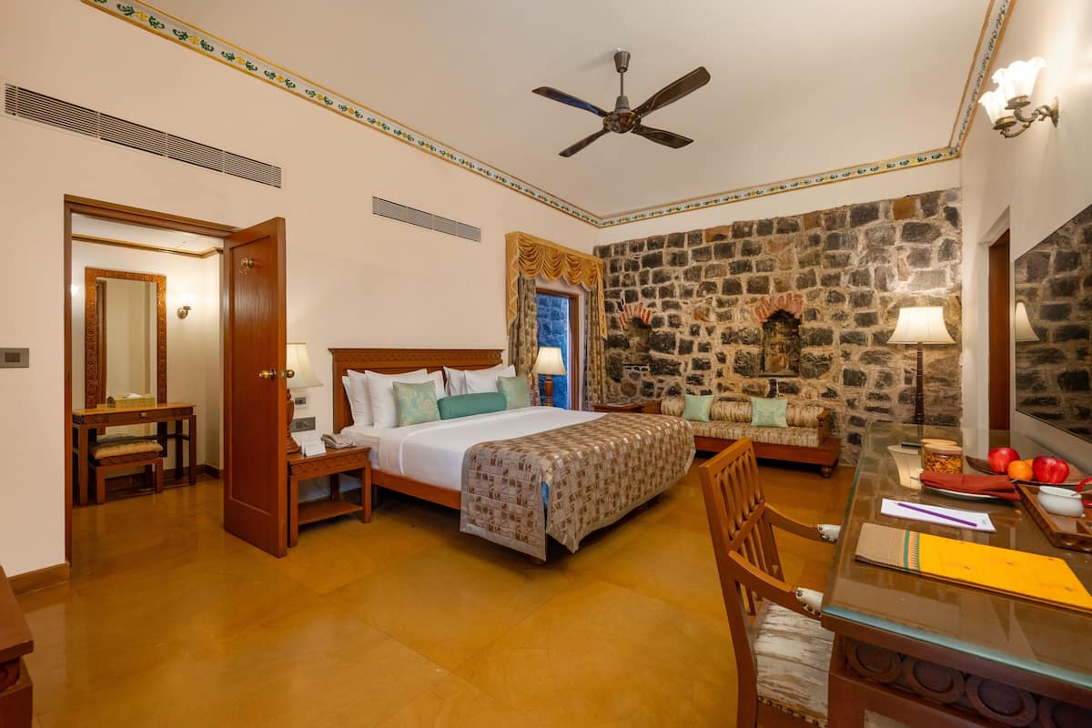 Une chambre d’hôtel avec un lit, un bureau, une chaise, un ventilateur de plafond et un mur en pierre.