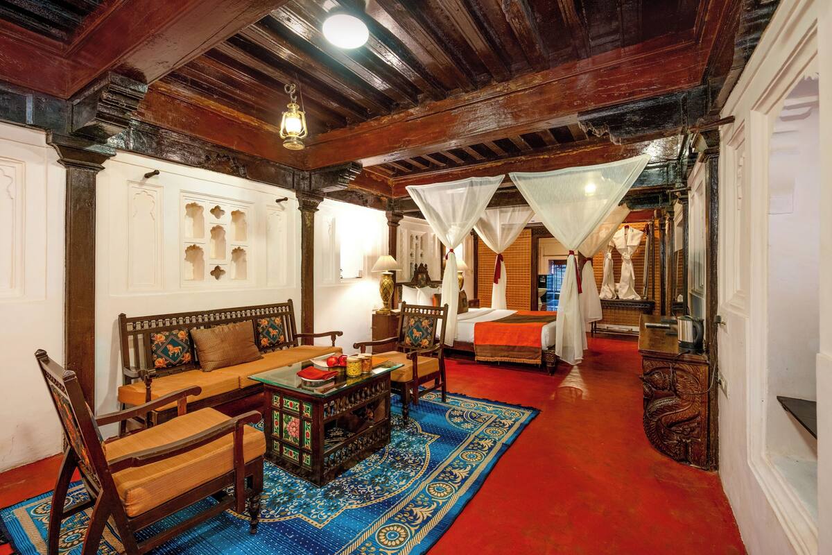Une chambre traditionnelle avec un plafond en bois, un lit recouvert d’une courtepointe rouge, un tapis bleu à motifs et une table en bois avec des chaises.