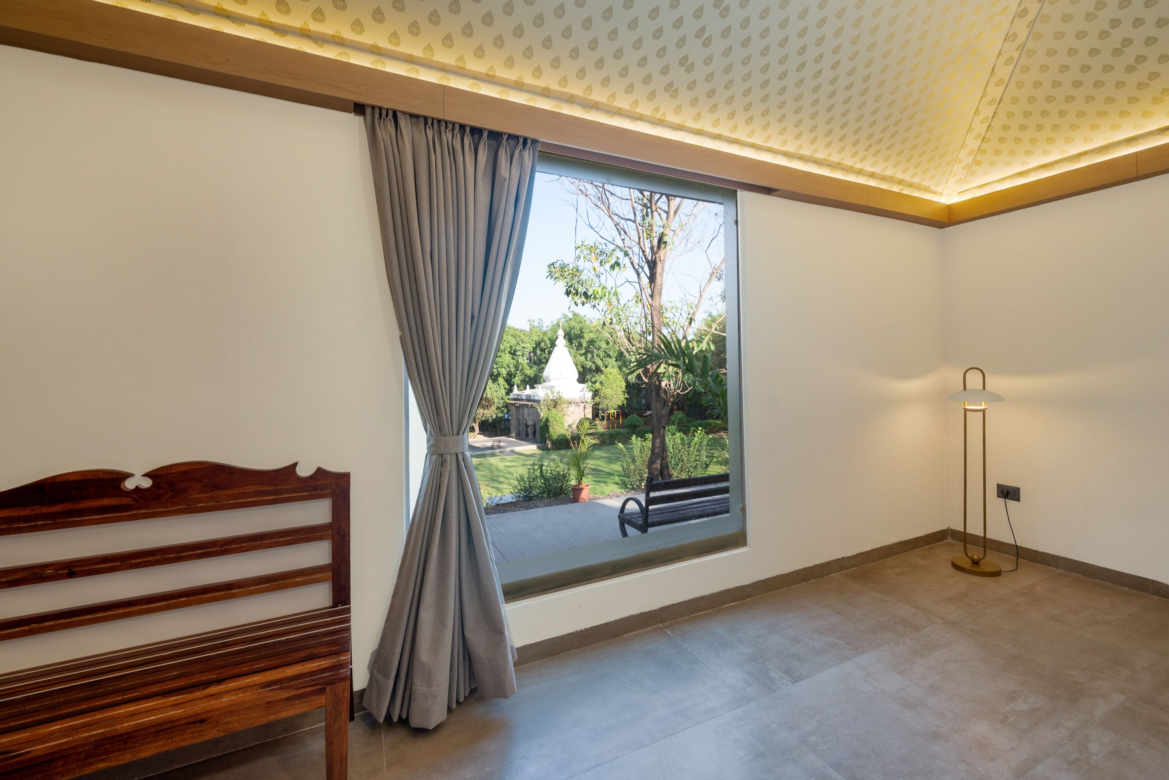 Ein Zimmer mit großem Fenster, einer Holzbank, einer Stehlampe und einer gemusterten Decke.