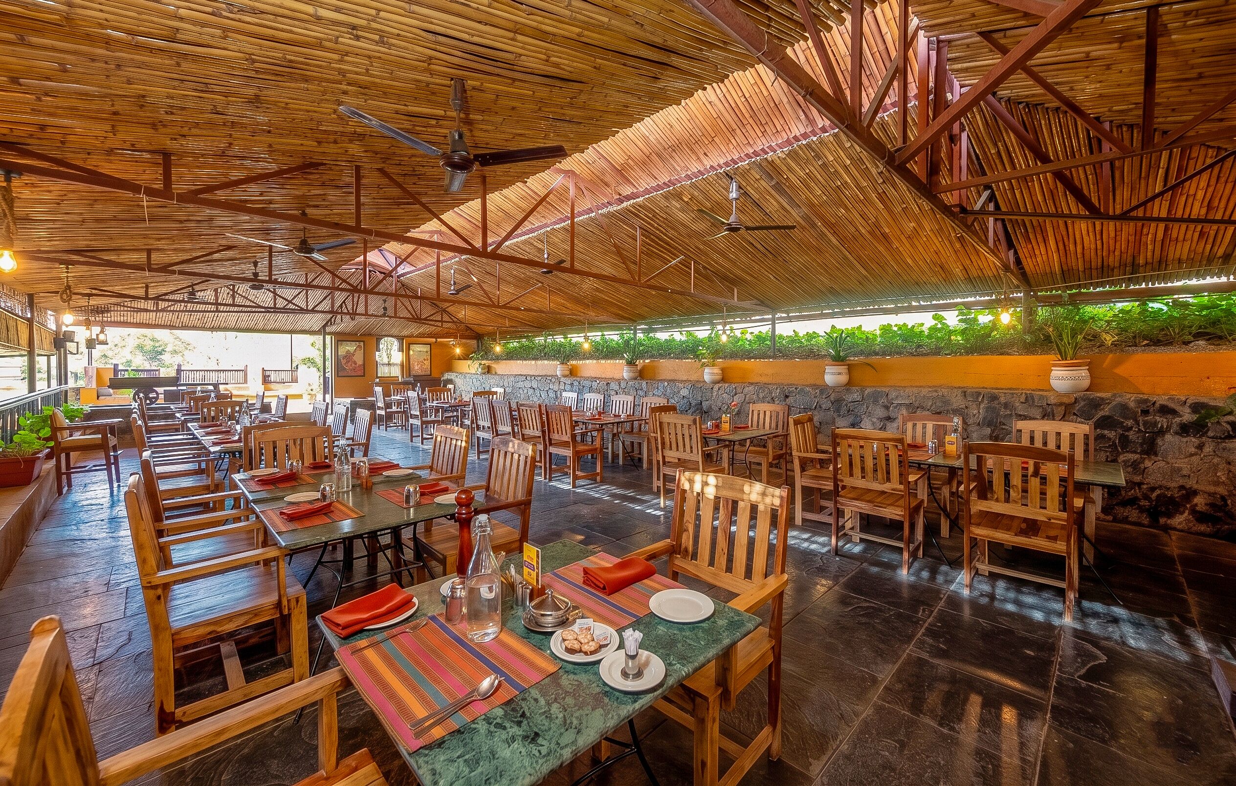 Un vaste espace de restauration intérieur, avec des tables et des chaises en bois, un plafond haut orné de poutres en bambou et un mur de pierre agrémenté de plantes.