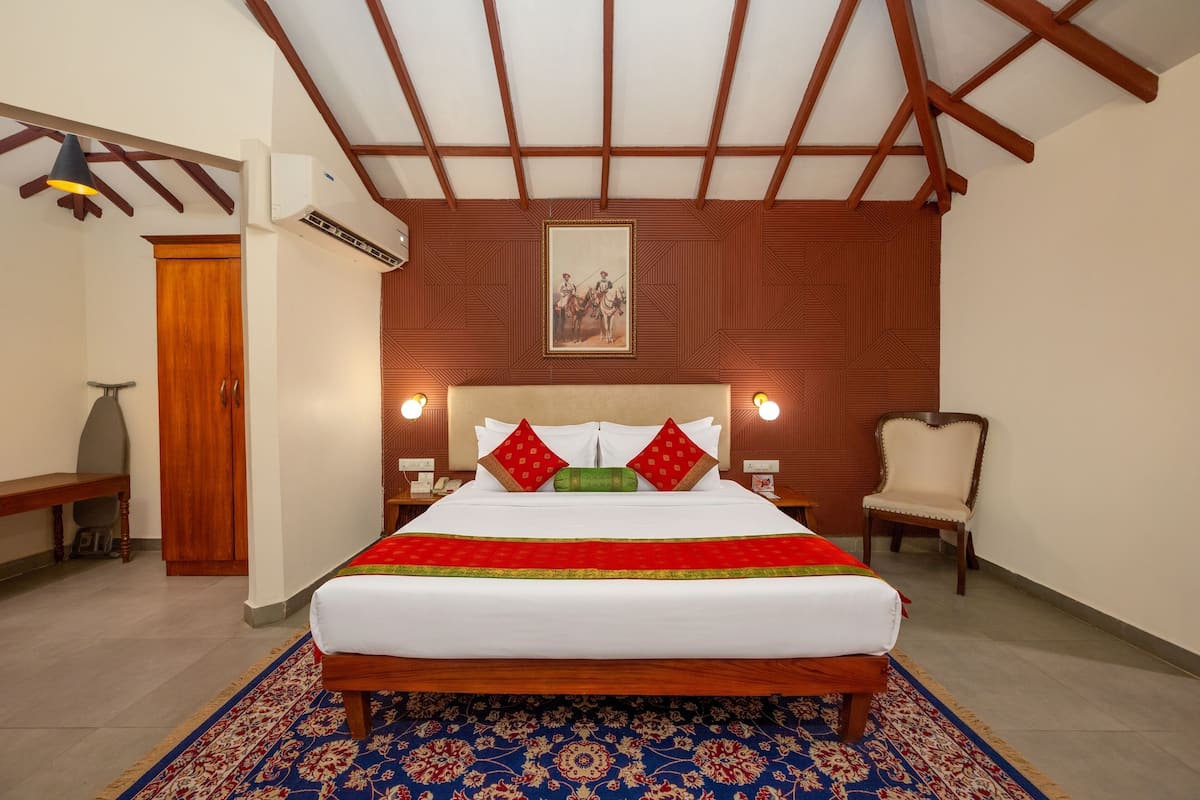 Une chambre d’hôtel avec un grand lit, une tête de lit en bois, un tapis à motifs, un banc en bois, une chaise et un climatiseur fixé au mur.