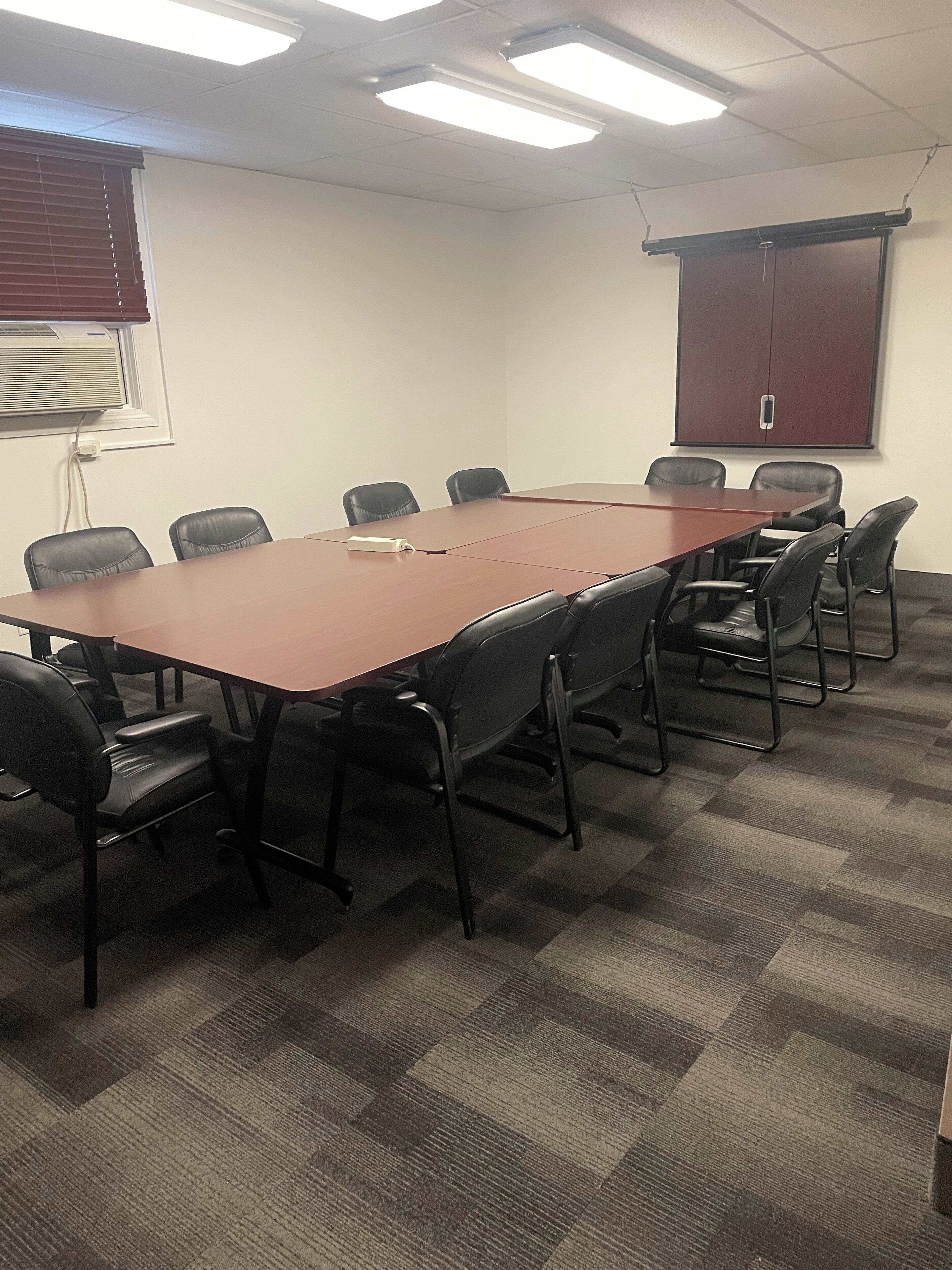 A conference room with a long table, black chairs, carpeted floor, ceiling lights, and a projector screen on the wall.