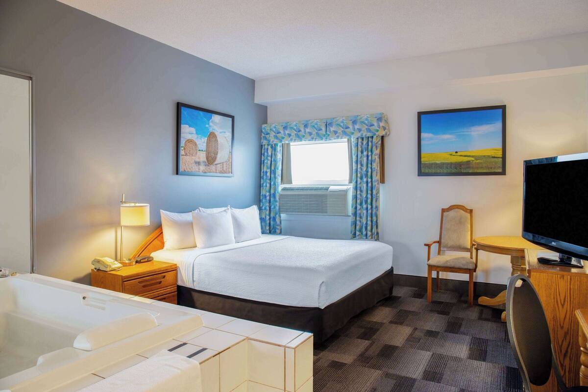 Ein Hotelzimmer mit einem großen Bett, einem Schreibtisch mit Stuhl, einem Fernseher, einer Schreibtischlampe und einem Fenster mit Vorhängen.