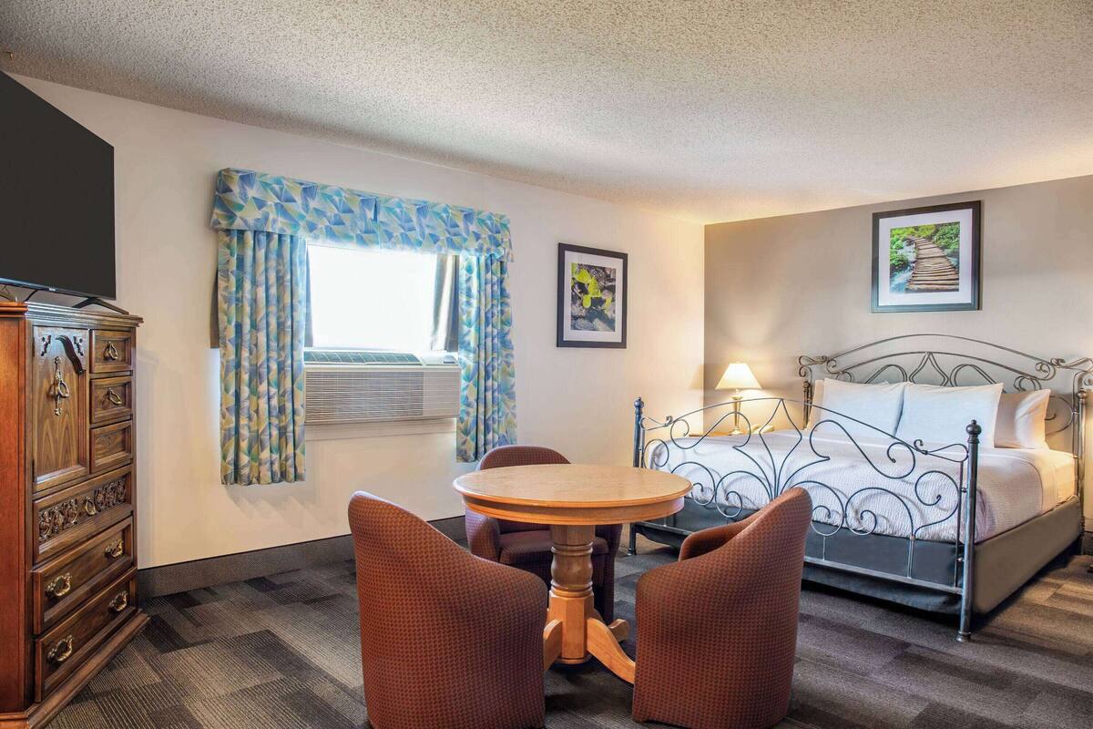 Ein Hotelzimmer mit einem Bett, einem runden Holztisch, zwei Stühlen, einer Kommode und einem Fenster mit Vorhängen.