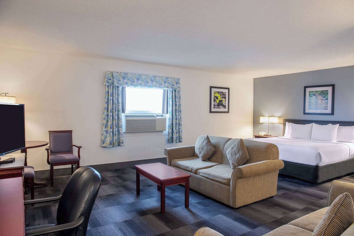 Ein Hotelzimmer mit Bett, einer Couch, einem Schreibtisch mit Stuhl, einem Fernseher und einem Fenster mit Vorhängen.