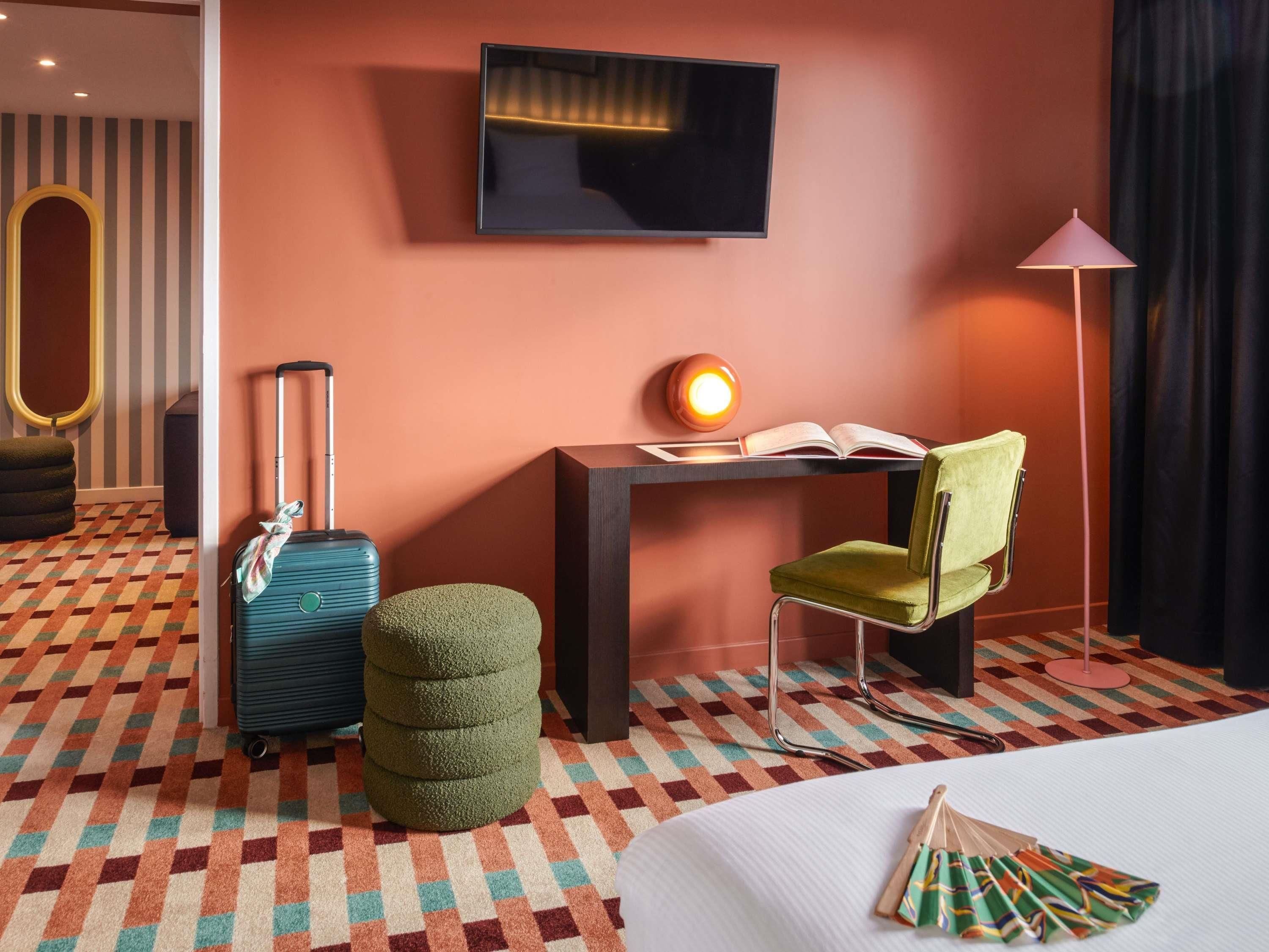 A hotel room with a bed, a desk with a lamp, a chair, a suitcase, and a small ottoman.
