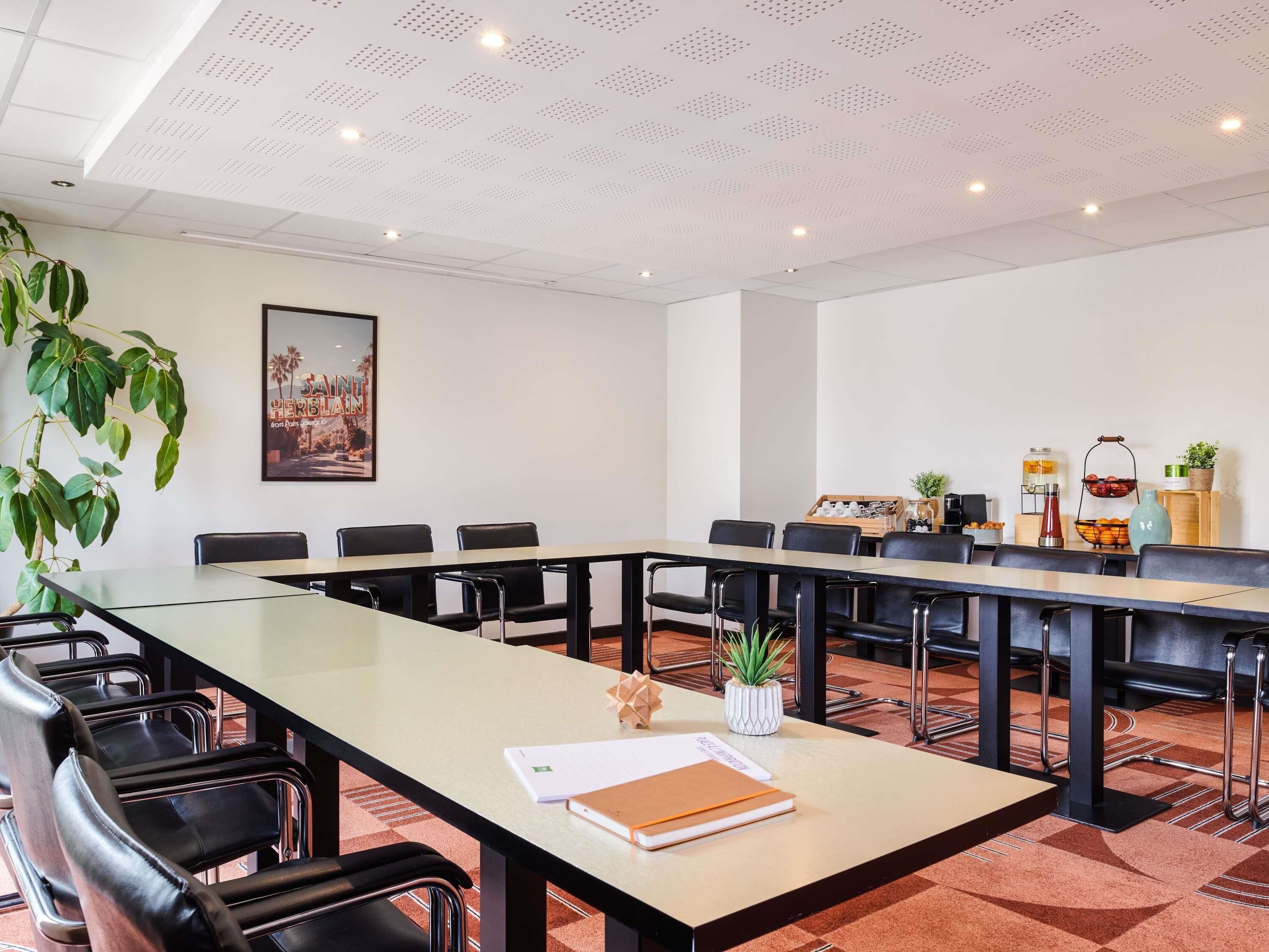 A conference room with a long table, black chairs, a potted plant, a poster, and a beverage station.