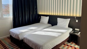Habitación de hotel con dos camas, una ventana con cortinas, un muro a rayas y una mesita de noche pequeña.