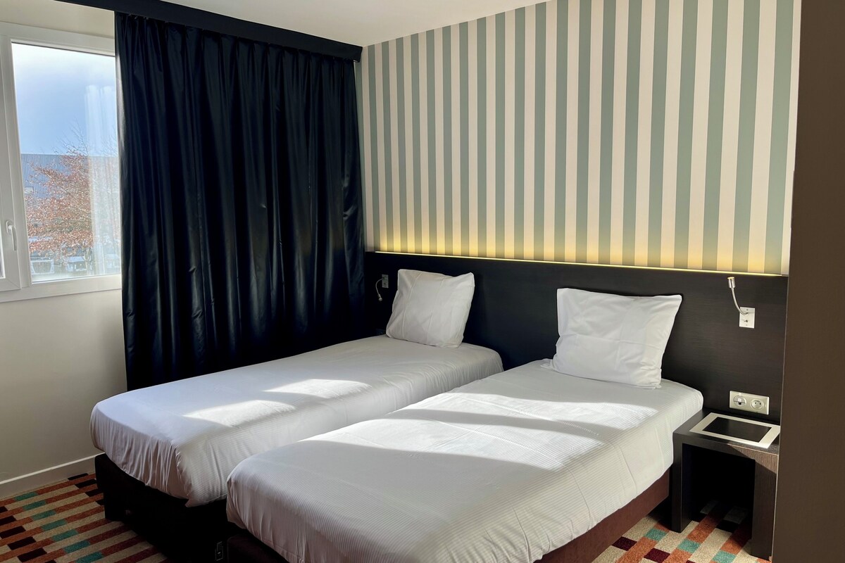 Habitación de hotel con dos camas, una ventana con cortinas, un muro a rayas y una mesita de noche pequeña.