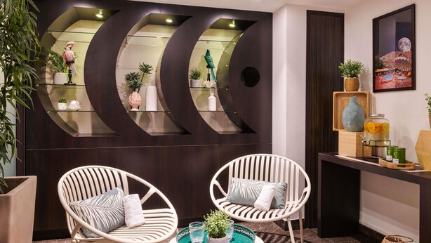 A modern interior with two white wicker chairs, a small round table with a green tray, and a dark wooden wall with glass shelves displaying decorative items. - ibis Styles Nantes Saint Herblain (Saint-Herblain)