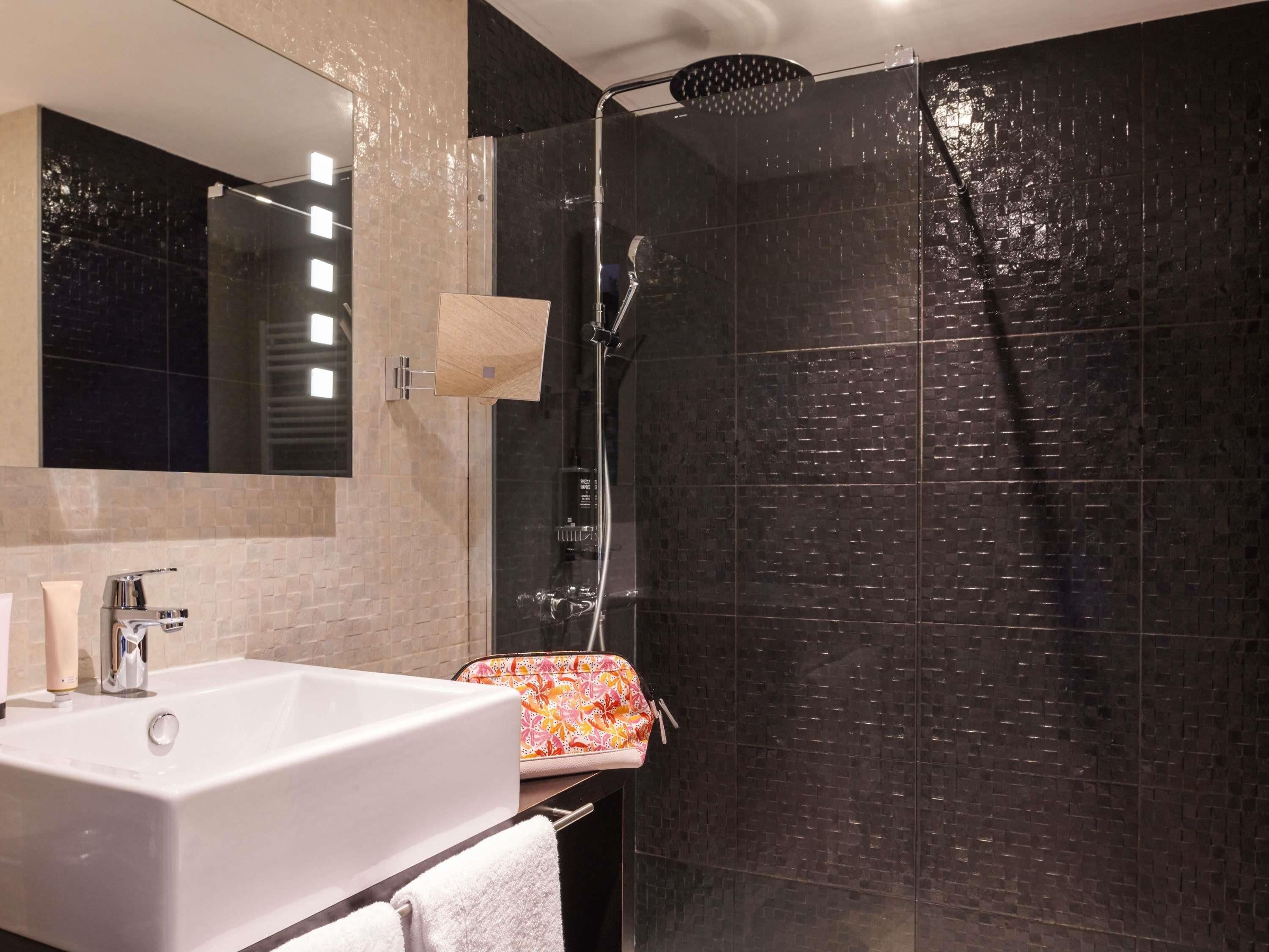 Une salle de bain moderne avec un grand miroir, une baignoire blanche, une douche à pommeau large et une serviette sur le sol.