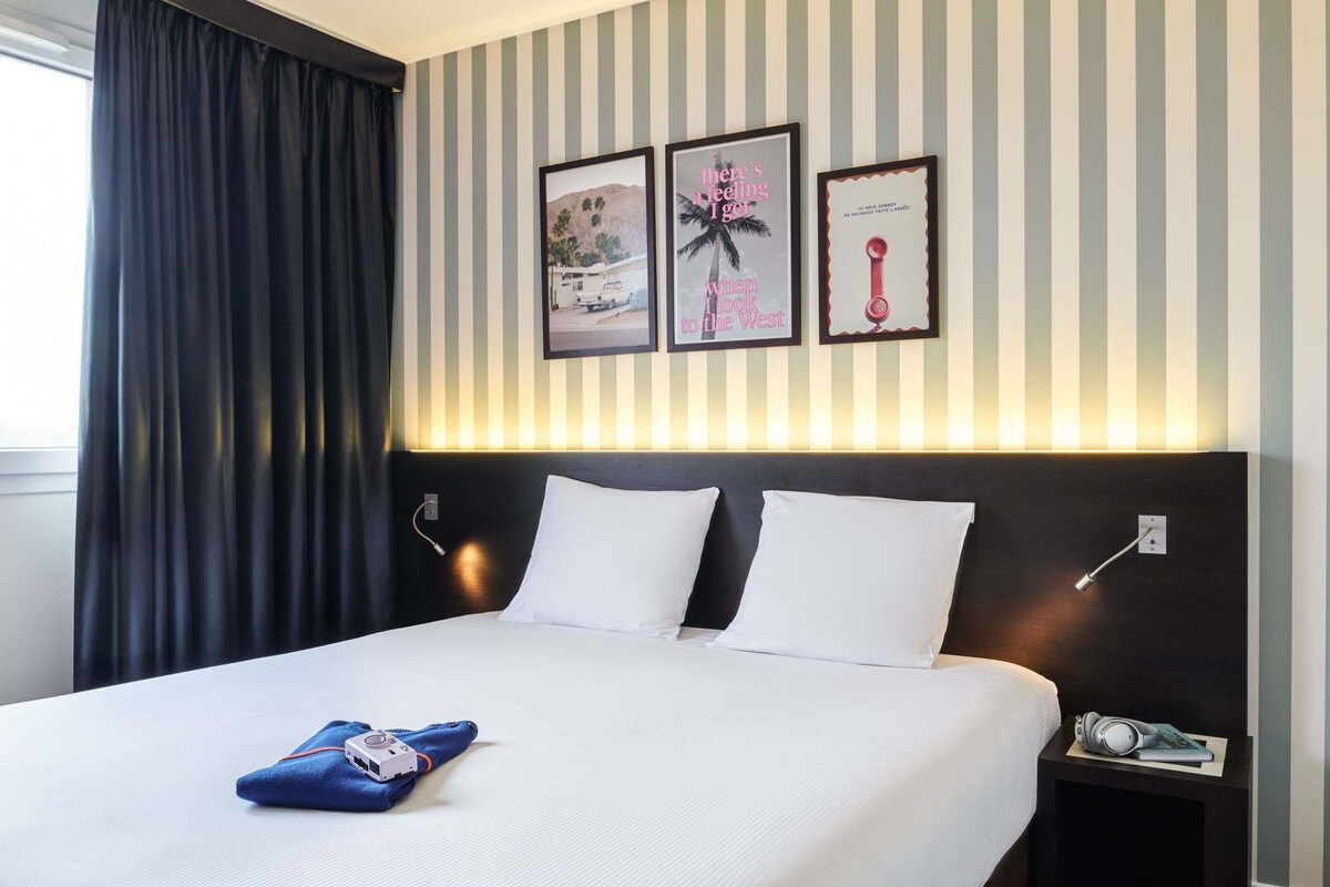 Una habitación de hotel con cama, mesita de noche y pared con papel tapiz a rayas y cuadros enmarcados.